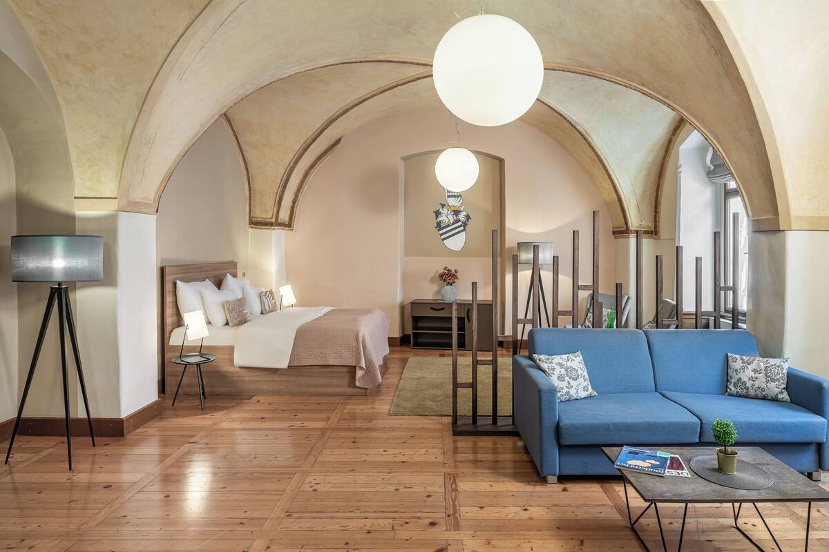 Ein Hotelzimmer mit Holzboden, einem Bett mit weißer Bettwäsche, einem blauen Sofa, einer Stehlampe und einer hohen, gewölbten Decke.