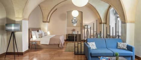 Habitación de hotel con piso de madera, cama con ropa blanca, sofá azul, lámpara de pie y un techo abovedado grande.