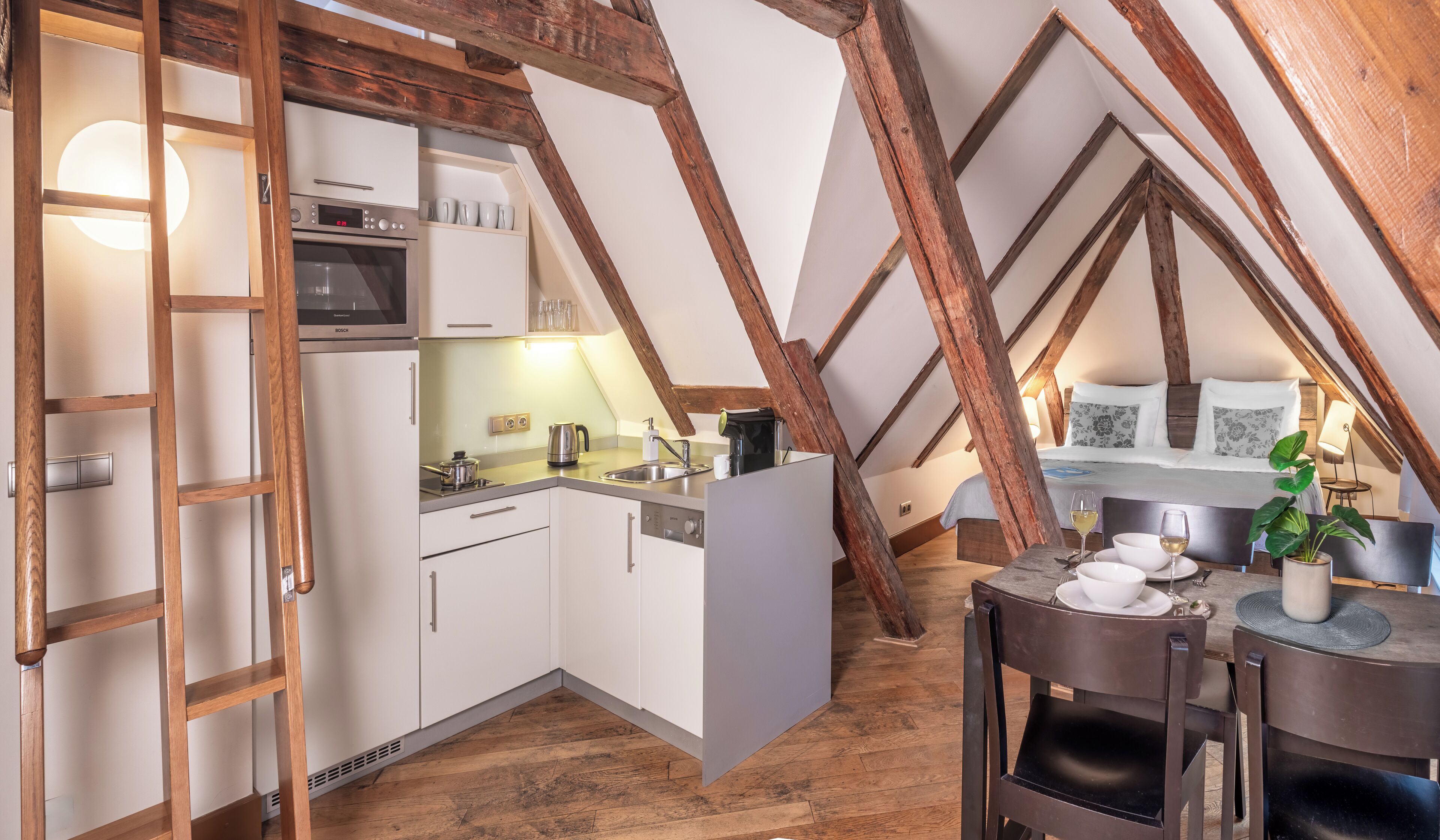 Une cuisine compacte avec des meubles blancs, un four Bosch et un évier. On aperçoit un coin repas avec une table et des chaises, ainsi qu'un lit à l'arrière-plan.