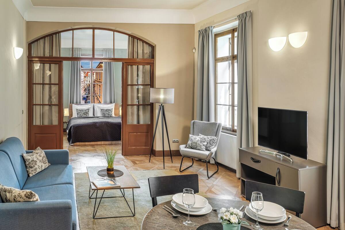 Ein Hotelzimmer mit einem Bett, einer Couch, einem Esstisch mit Stühlen, einem Fernseher und einem großen Fenster mit Vorhängen.