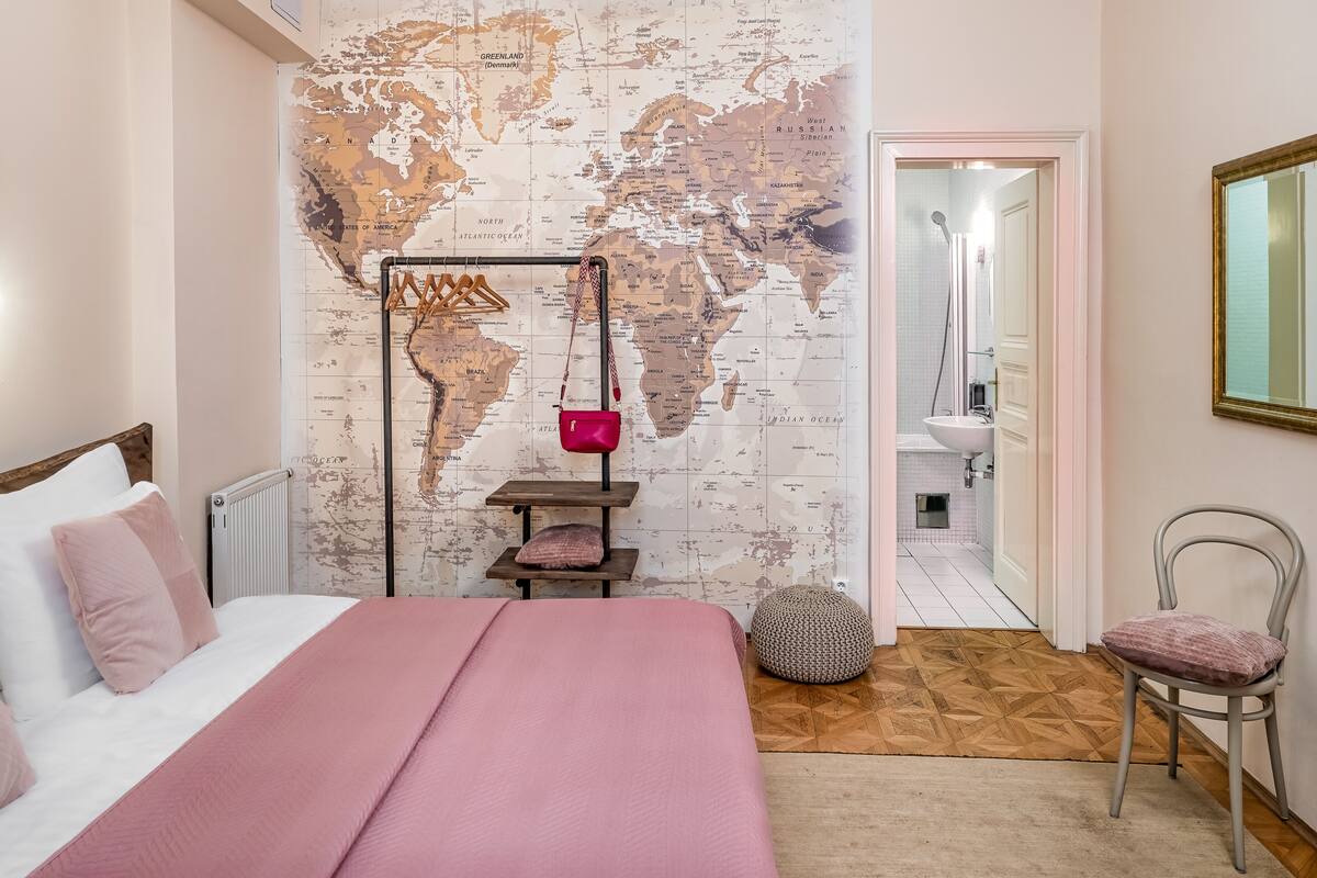 Ein Schlafzimmer mit einer Weltkarte an der Wand, einem Bett mit rosa Bettwäsche, einem kleinen Tisch mit einer pinkfarbenen Tasche und einem Stuhl.