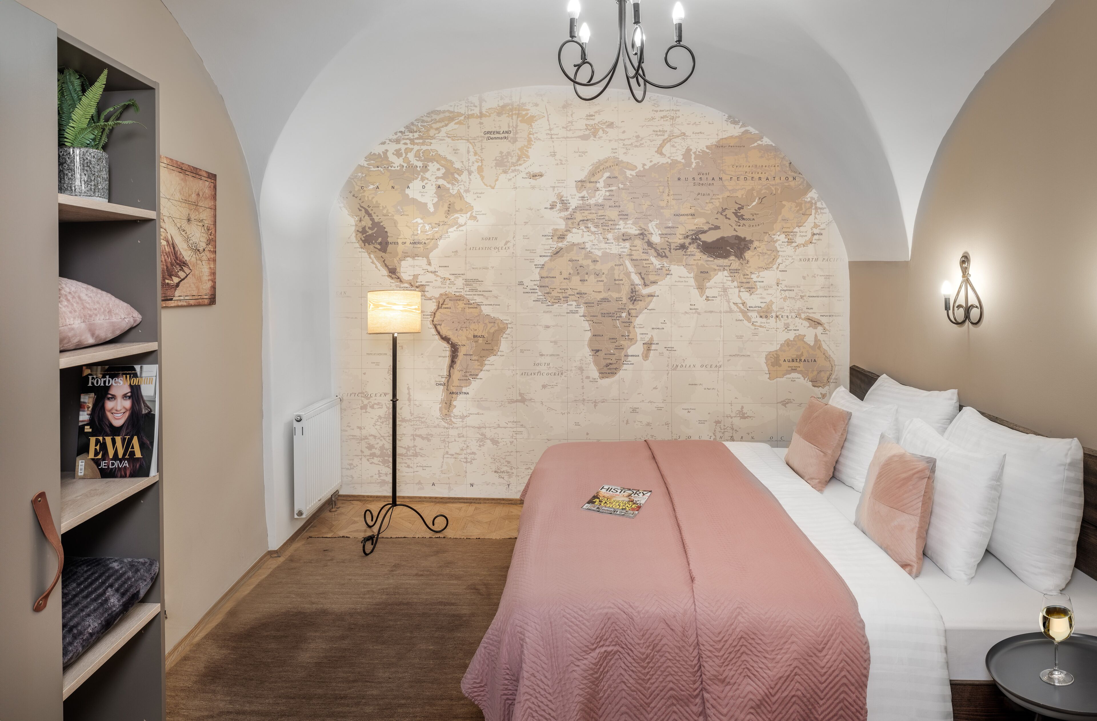 A bedroom with a world map wall, a bed with white and pink bedding, a small table with a glass, and a bookshelf with magazines.