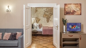Una habitación con un sofá gris, una cama con manta rosa, un mapa del mundo en la pared, un televisor y una planta sobre un mueble.