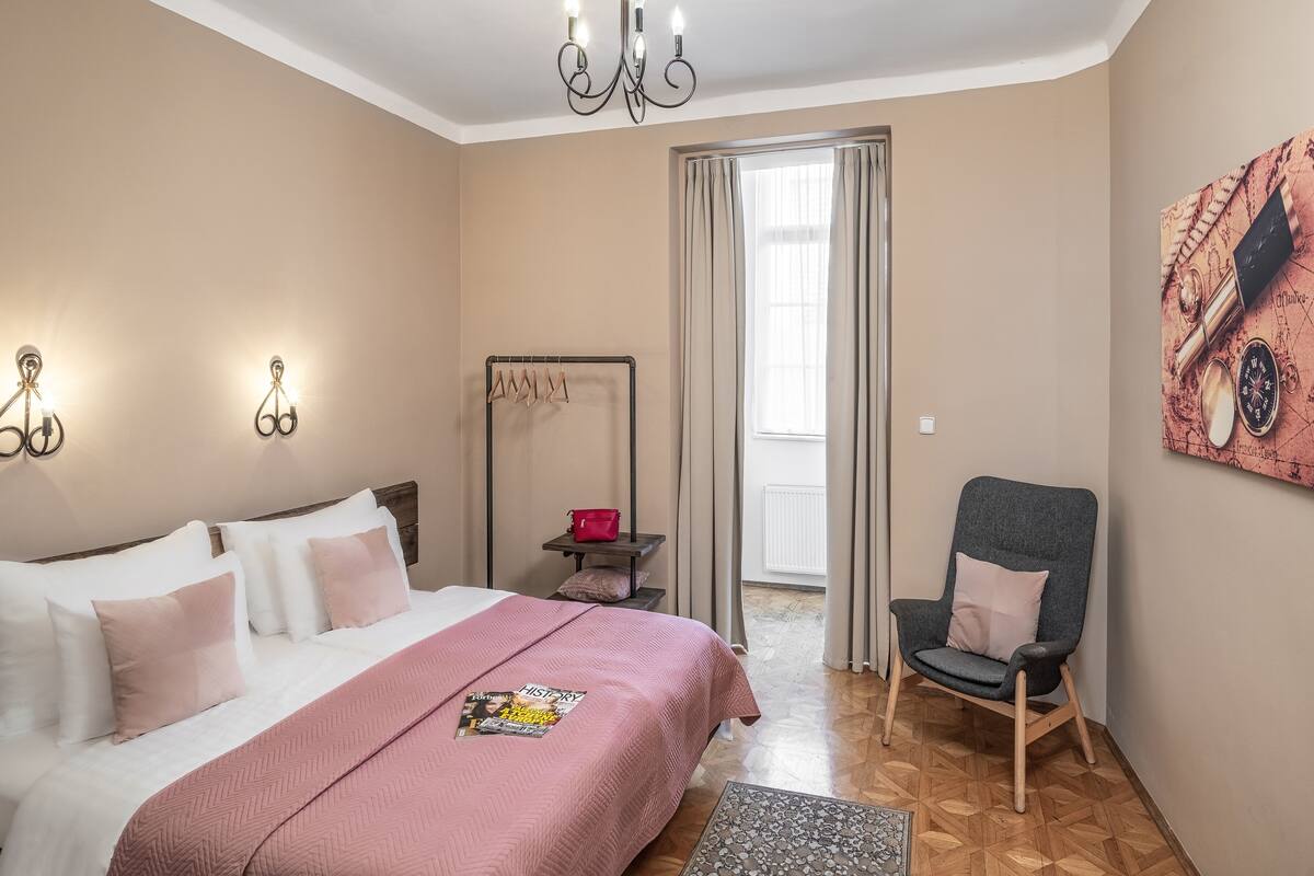 Ein Schlafzimmer mit einem Bett, einem Sessel, einem Wandhaken zur Kleideraufhängung und einem Fenster mit Vorhängen.