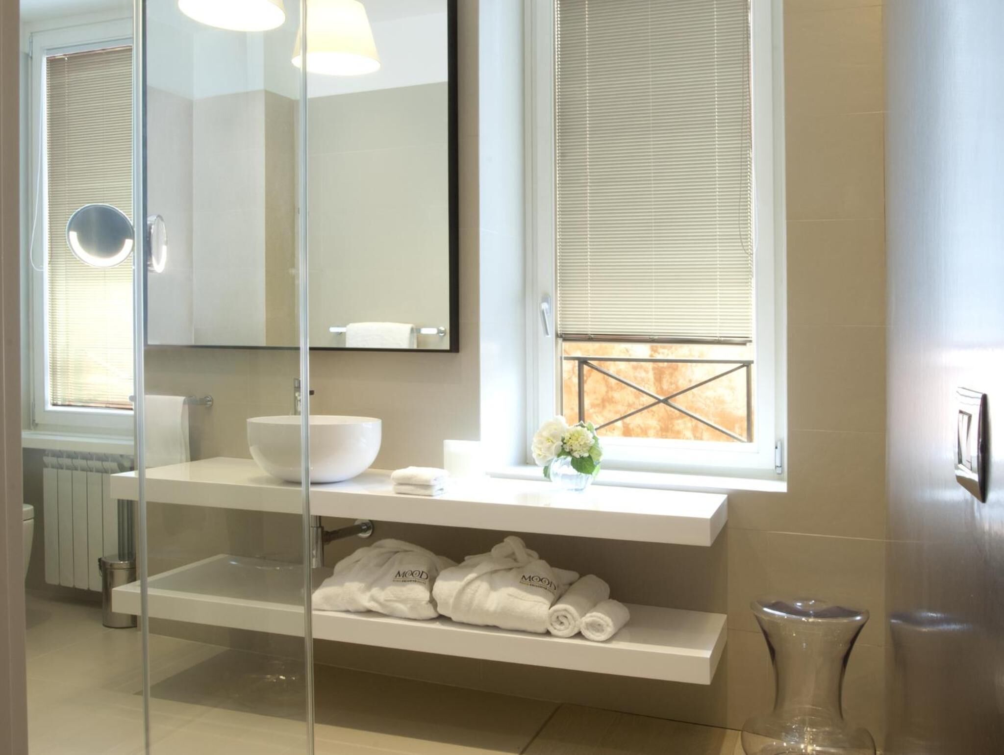 Une salle de bain moderne avec une douche à parois de verre, un grand miroir, un lavabo blanc et une fenêtre avec des stores.