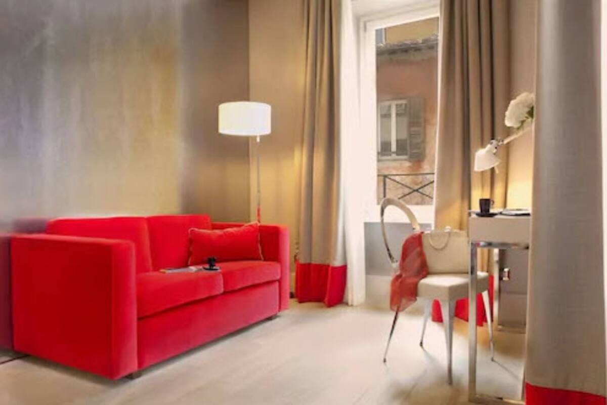 A modern hotel room with a red sofa, a desk with a lamp, and a view of the city through large windows.