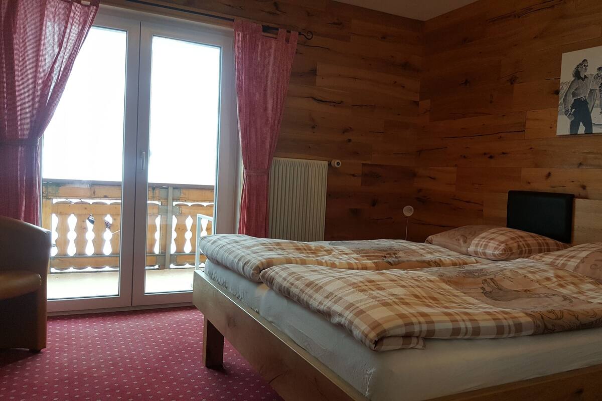 A wooden room with a bed, a window with curtains, a painting on the wall, and a balcony with a railing.