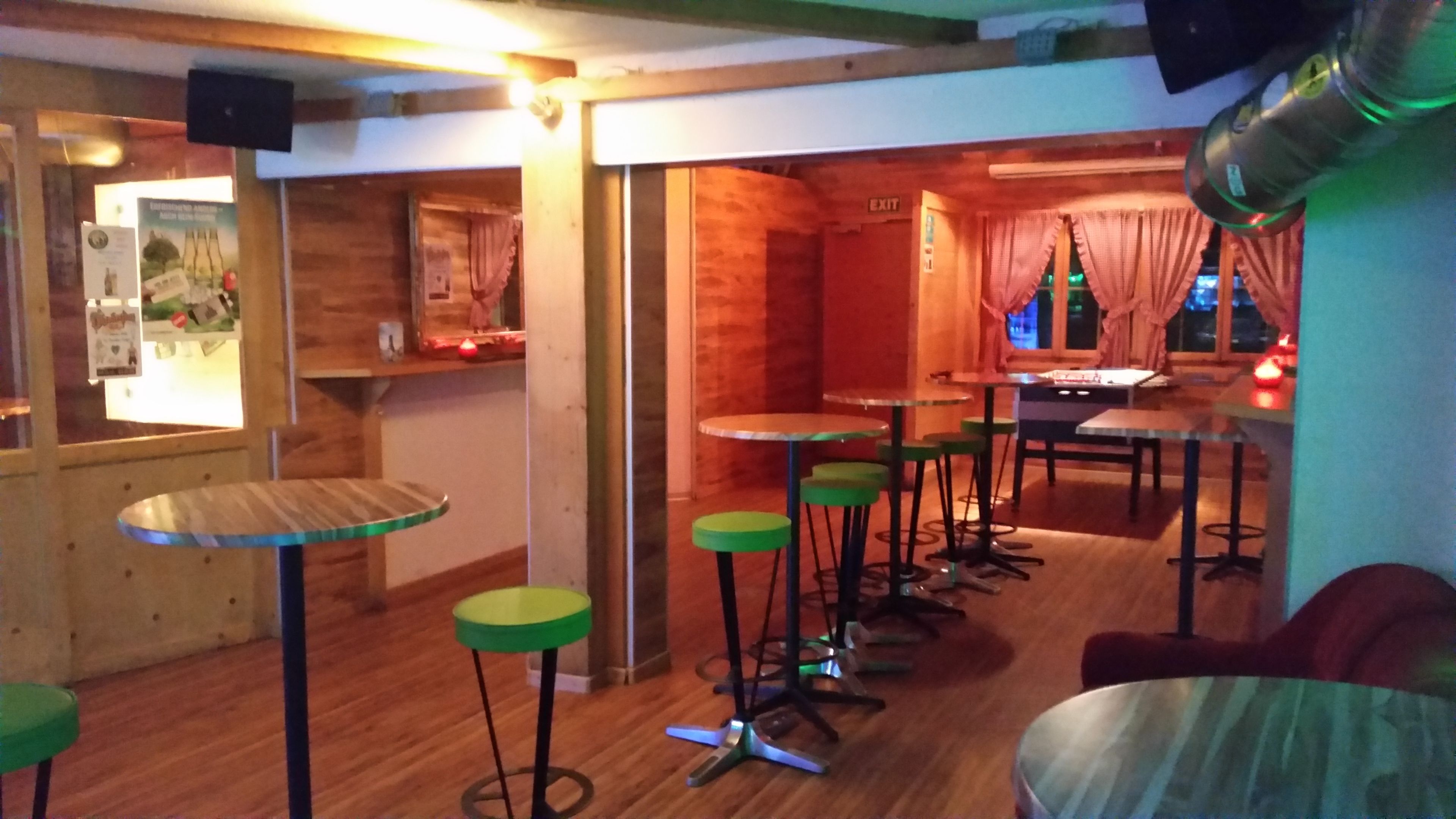 A cozy bar area with wooden walls, green stools, and a mix of round and square tables.