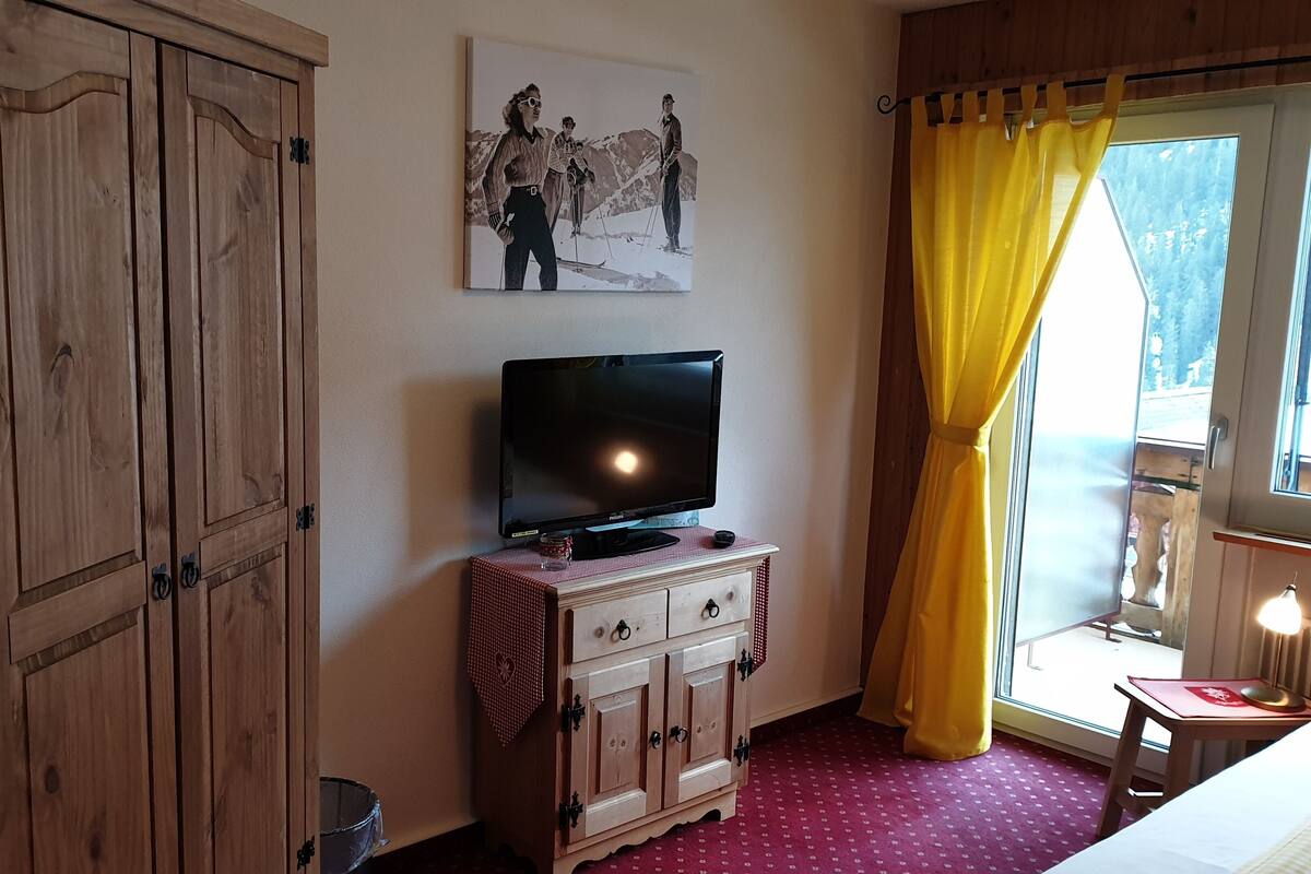 A room with wooden cabinets, a television on a small table, a window with yellow curtains, and a painting on the wall.