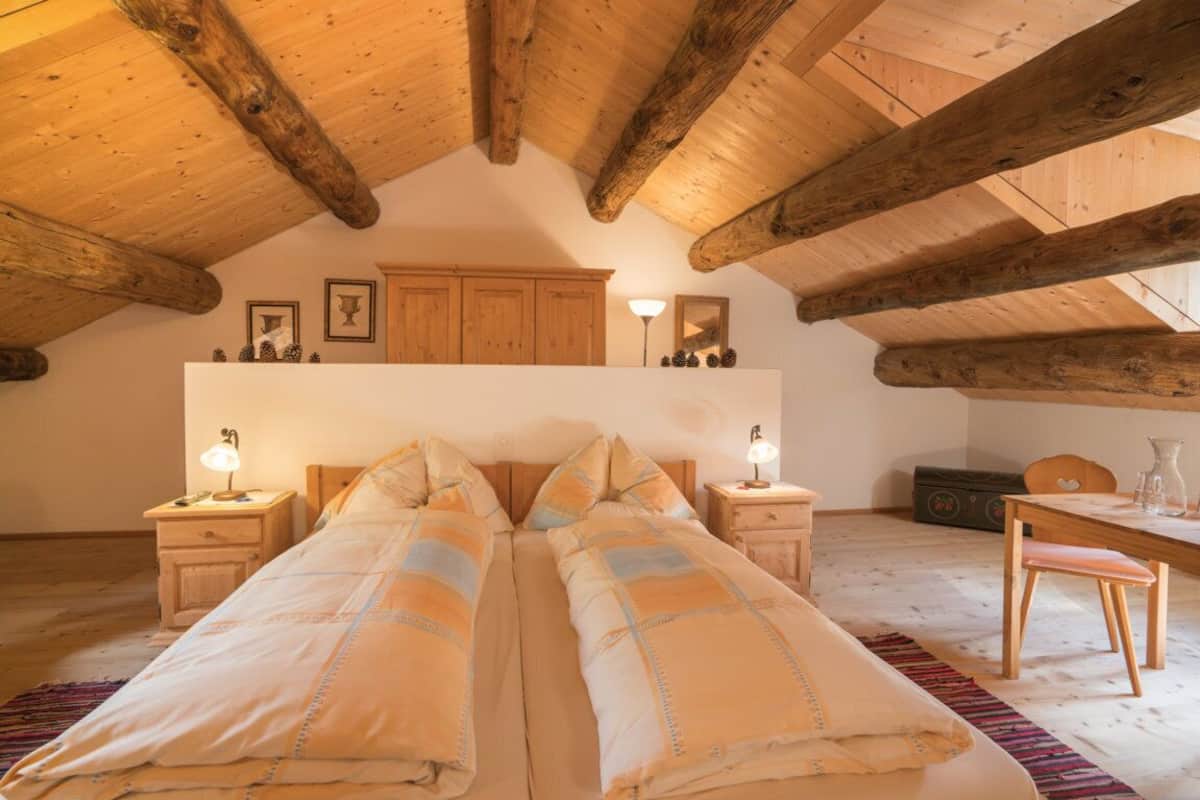 Ein gemütliches Schlafzimmer im Dachgeschoss mit freiliegenden Holzbalken, einem großen Bett mit passenden Kissen, Nachttischen mit Lampen und einem Holzschrank über dem Bett.