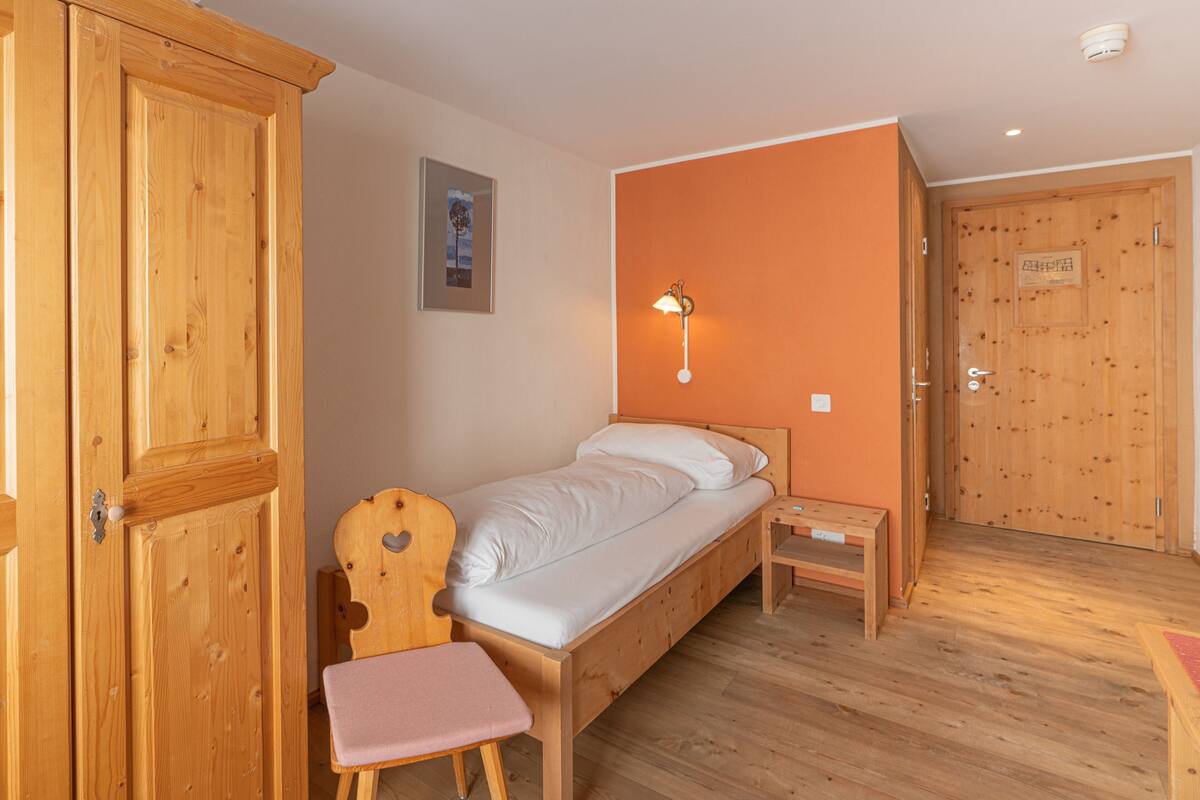 Ein Zimmer mit Holzboden, einem Einzelbett, einem Nachttischchen, einem Sessel und einem Kleiderschrank.
