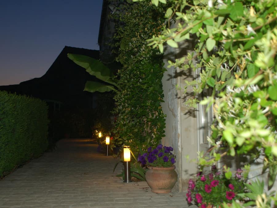 Un sendero bordeado por macetas con plantas y faroles iluminados, que conduce a un edificio con techo inclinado.