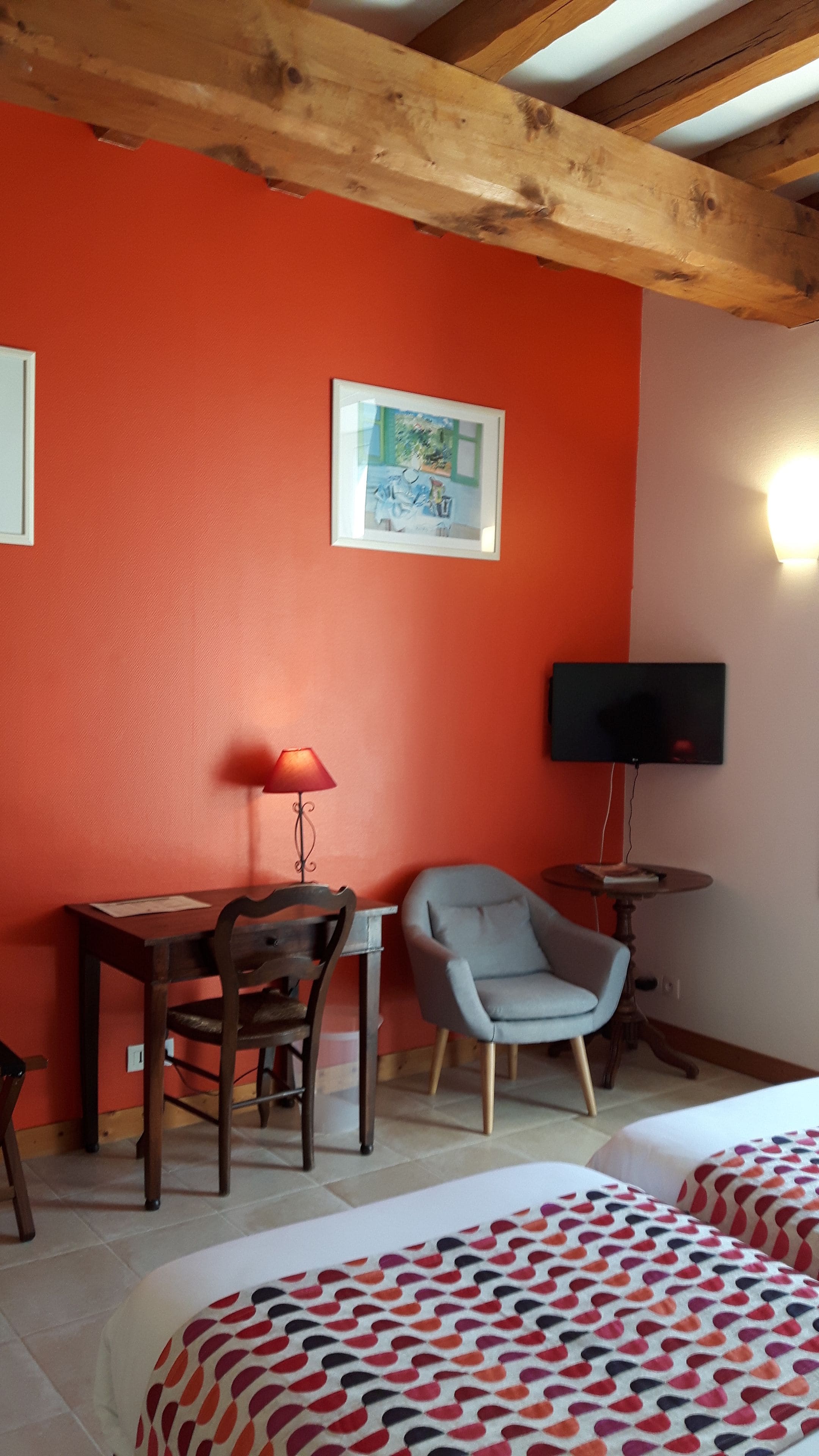 A room with a wooden ceiling, a red wall, a framed picture, a television, a desk with a chair, and a bed with patterned bedding.