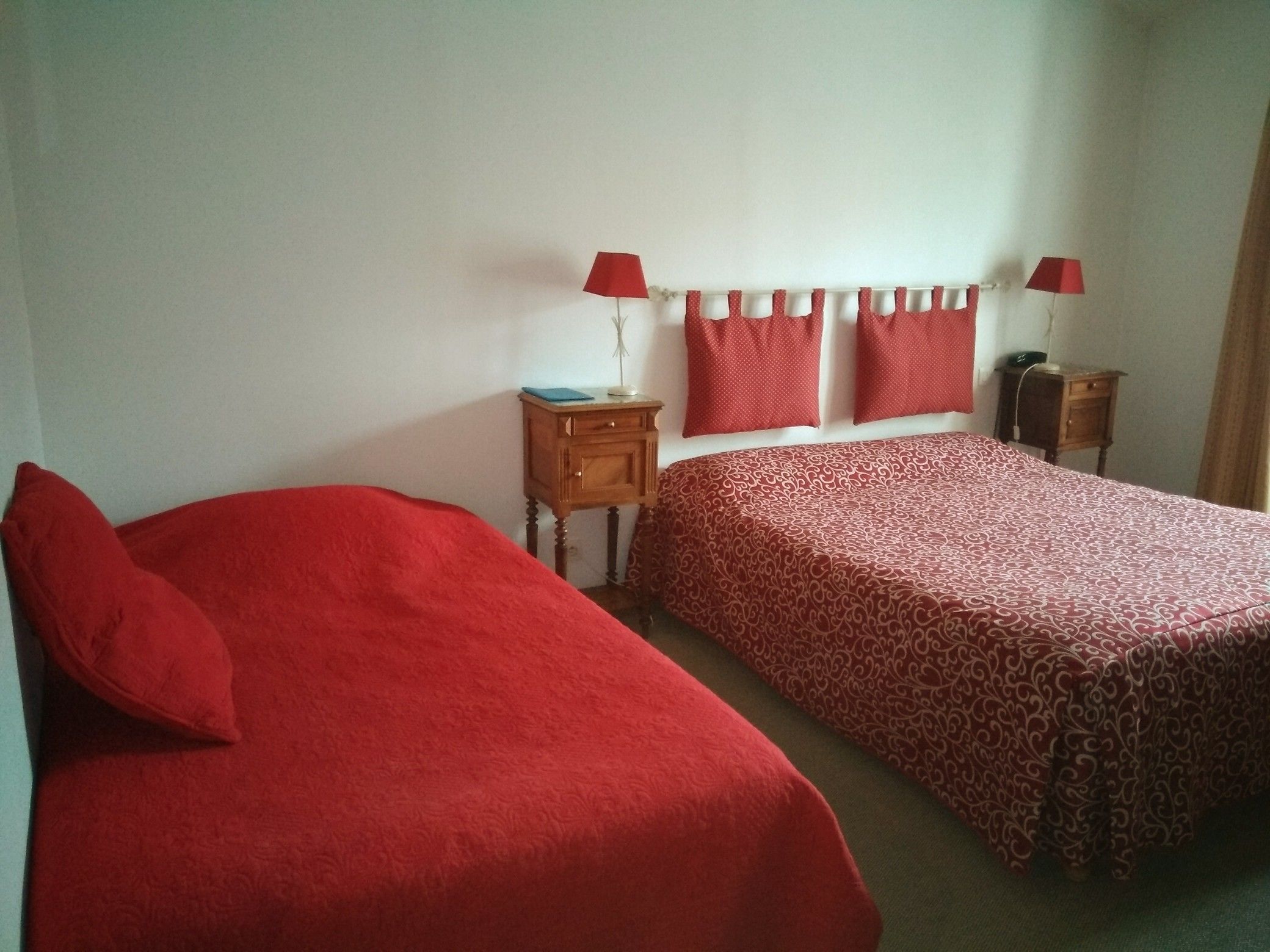 A room with two single beds, each with a red bedspread and matching pillows. There are two bedside tables with lamps, a small wooden nightstand, and a wall-mounted shelf with a lamp.