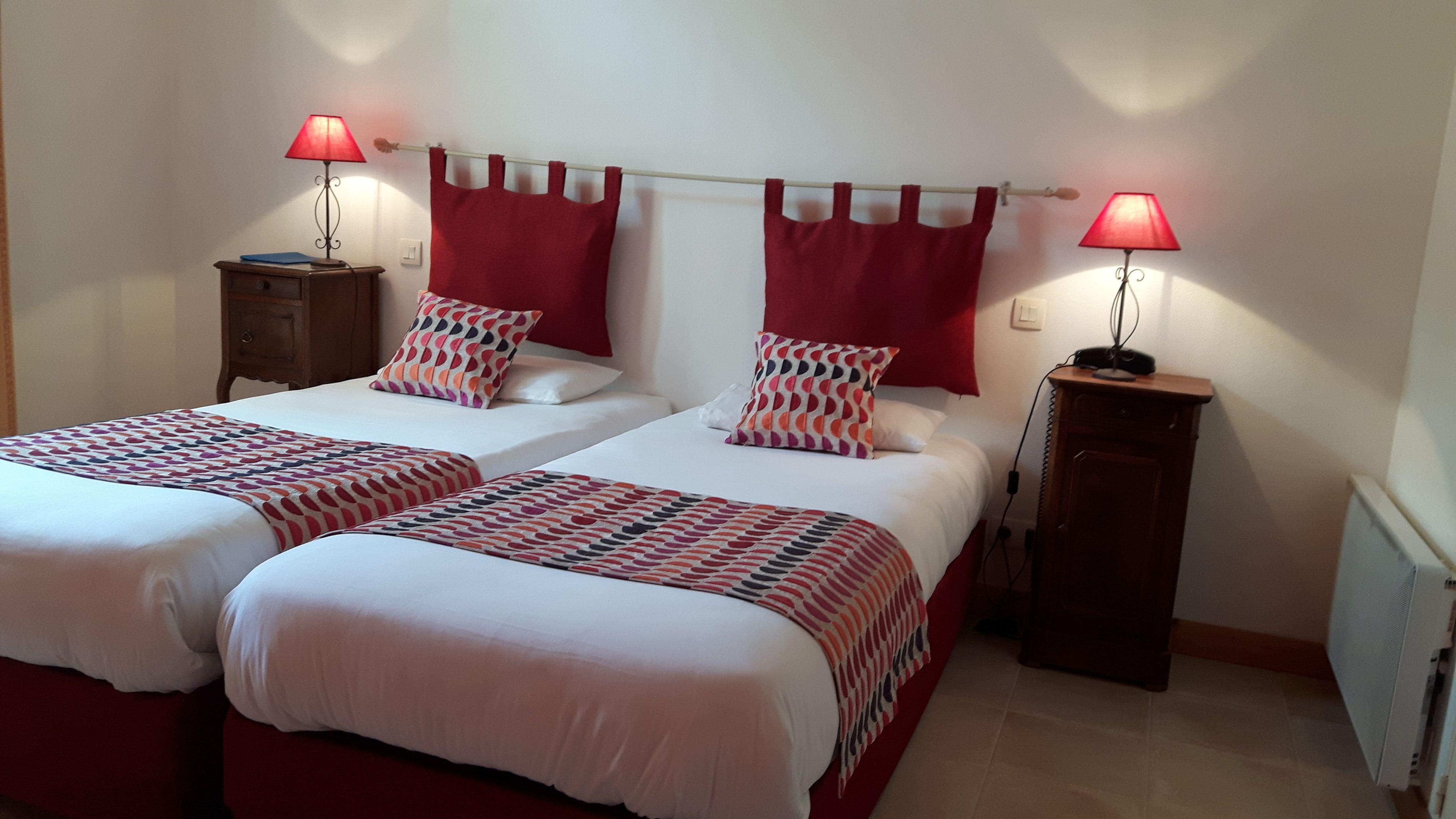 A double room with two beds, each with a red headboard, white bedding, and patterned throw blankets. There are two bedside tables with lamps, and a radiator on the wall.