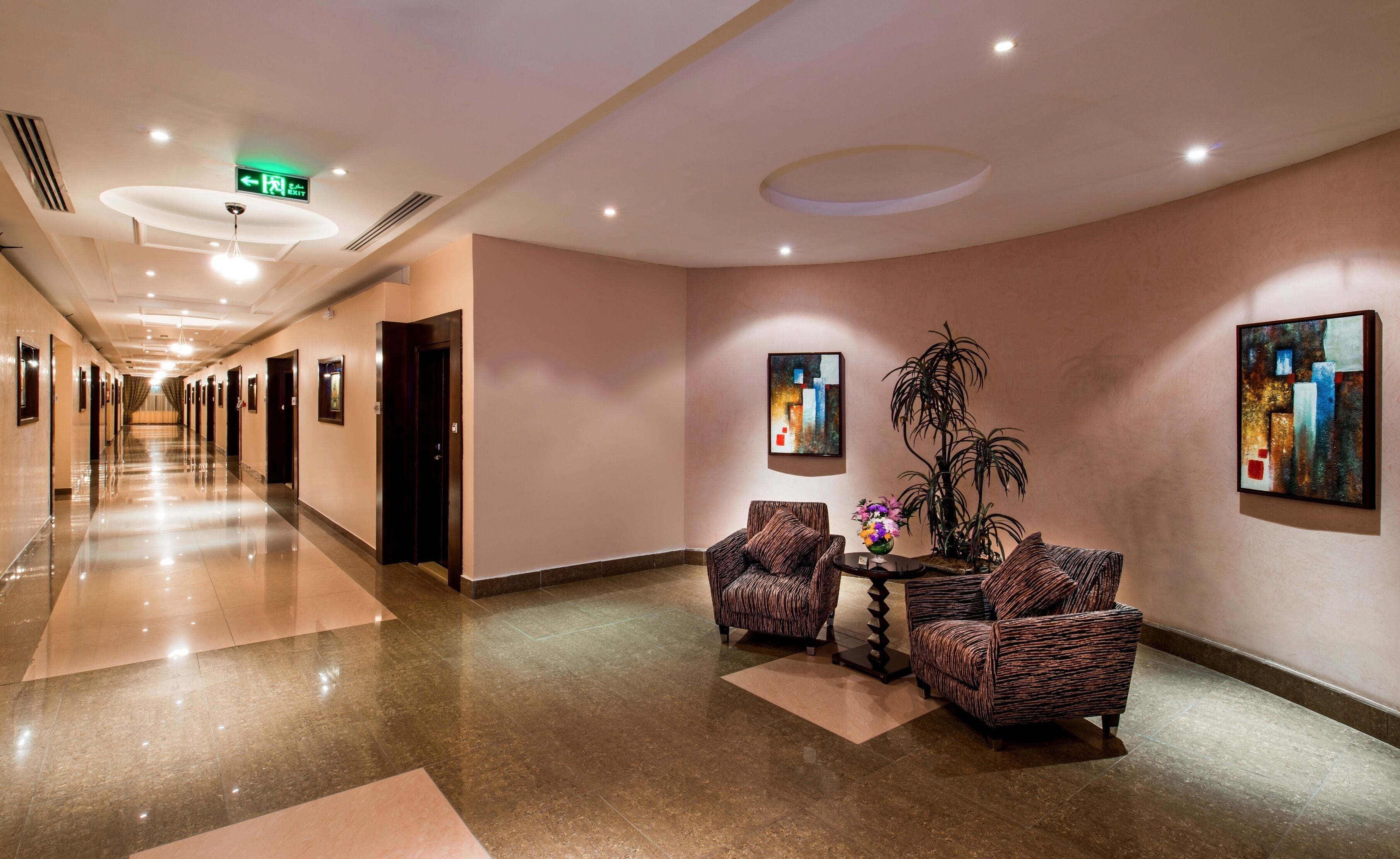 Un hall de hotel con un largo pasillo, dos sillones, una mesa con un florero y obras de arte en las paredes.