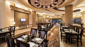 Ruime hotel eetzaal met meerdere tafels en stoelen, een aan de muur bevestigde televisie en een modern plafondontwerp.