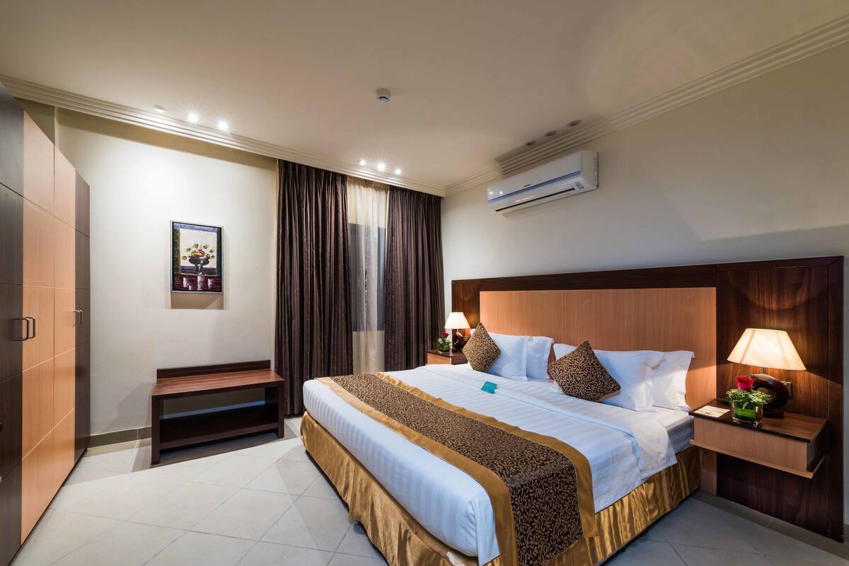 Habitación de hotel con una cama grande, un banco, una mesita de noche con lámpara y un equipo de aire acondicionado en la pared.