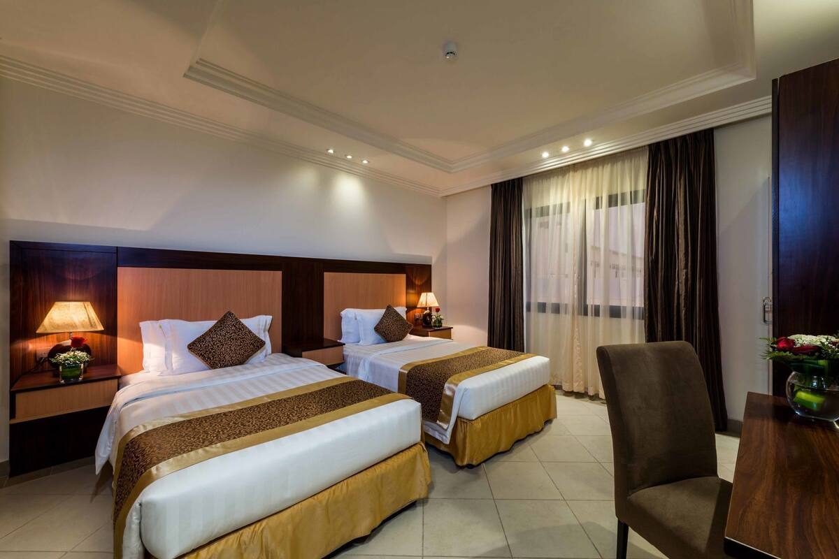 Habitación de hotel con dos camas, cabecero de madera, lámparas de noche, una silla y una mesa.