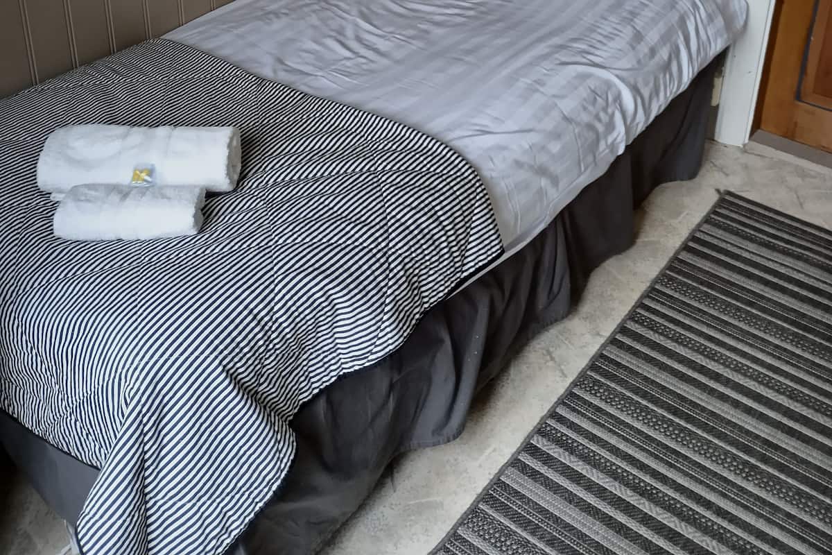 Una cama individual con una colcha a rayas, una toalla, una lámpara de pared y un cuadro en la pared.