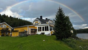A large yellow house with a white porch and a garage, surrounded by a grassy yard and a lake. - Lugnet Malung Hotel (Malung)