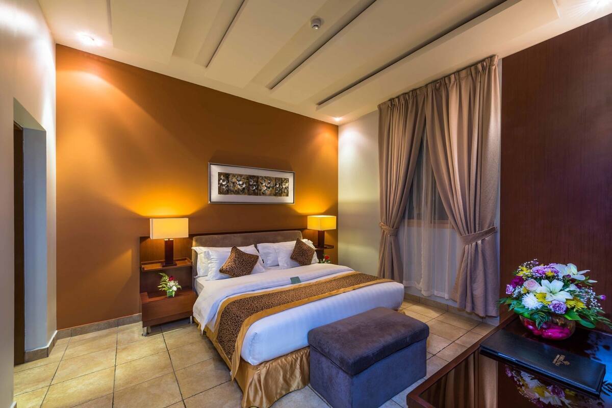 Une chambre d’hôtel avec un grand lit, des lampes de chevet, une table de nuit, un bureau agrémenté d’un vase de fleurs et une fenêtre avec des rideaux.