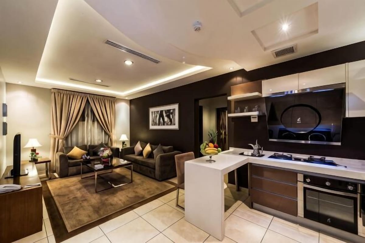 A modern living room with a sofa, coffee table, and TV. There's a kitchen with a stove and oven.