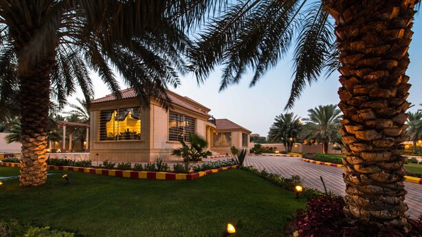 A building with a tiled roof and large windows, surrounded by well-maintained grass and palm trees. - Boudl Nakhil  (Unaizah)