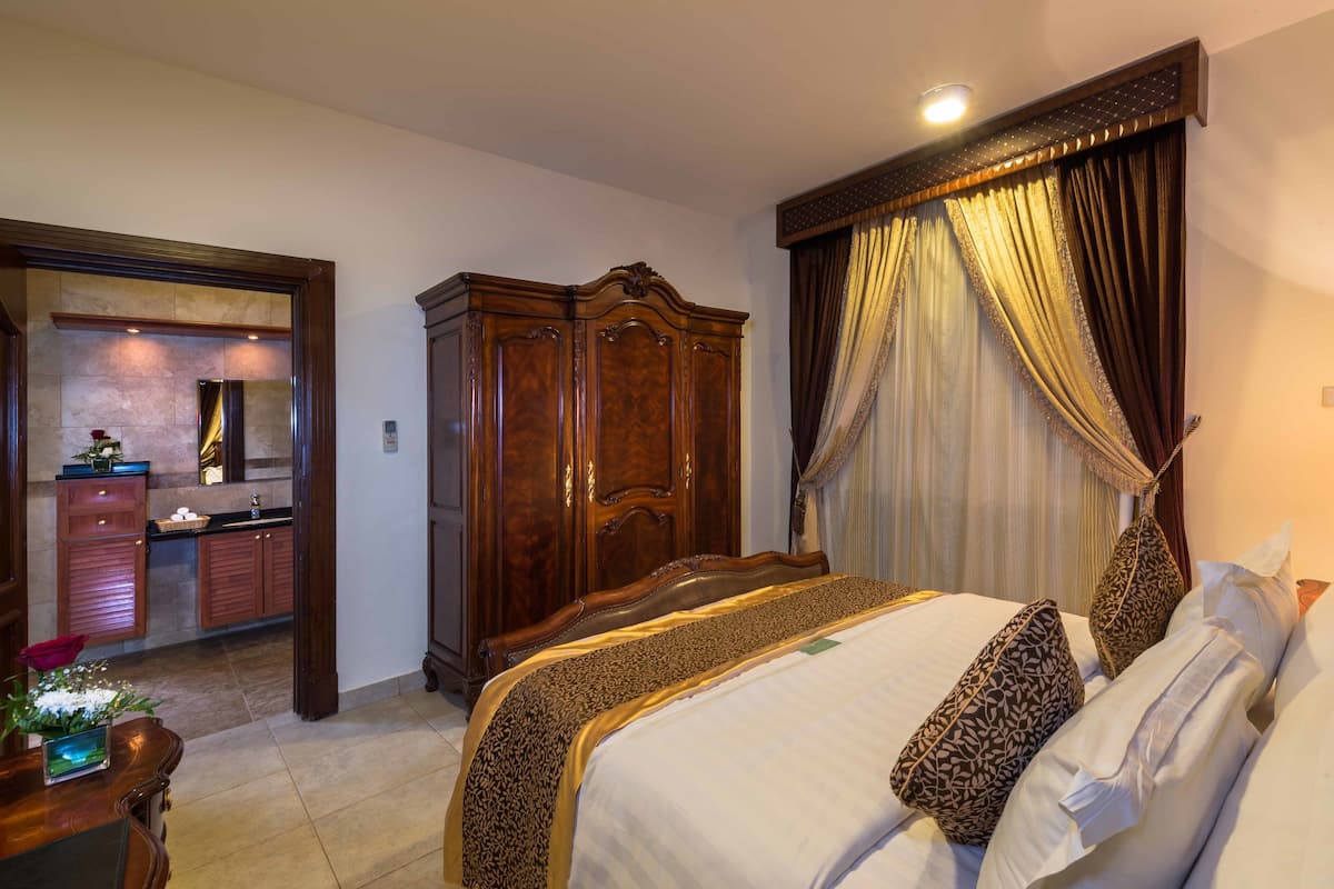 Quarto de hotel com uma cama grande, um guarda-roupa, uma secretária com abajur e um banheiro visível através de uma porta aberta.