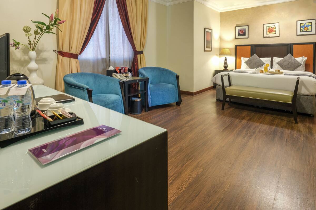 A hotel room with a bed, two blue armchairs, a desk with a water bottle, a vase with flowers, and a bedside table with a lamp.