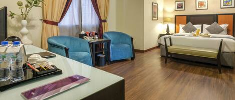 A hotel room with a bed, two blue armchairs, a desk with a water bottle, a vase with flowers, and a bedside table with a lamp.