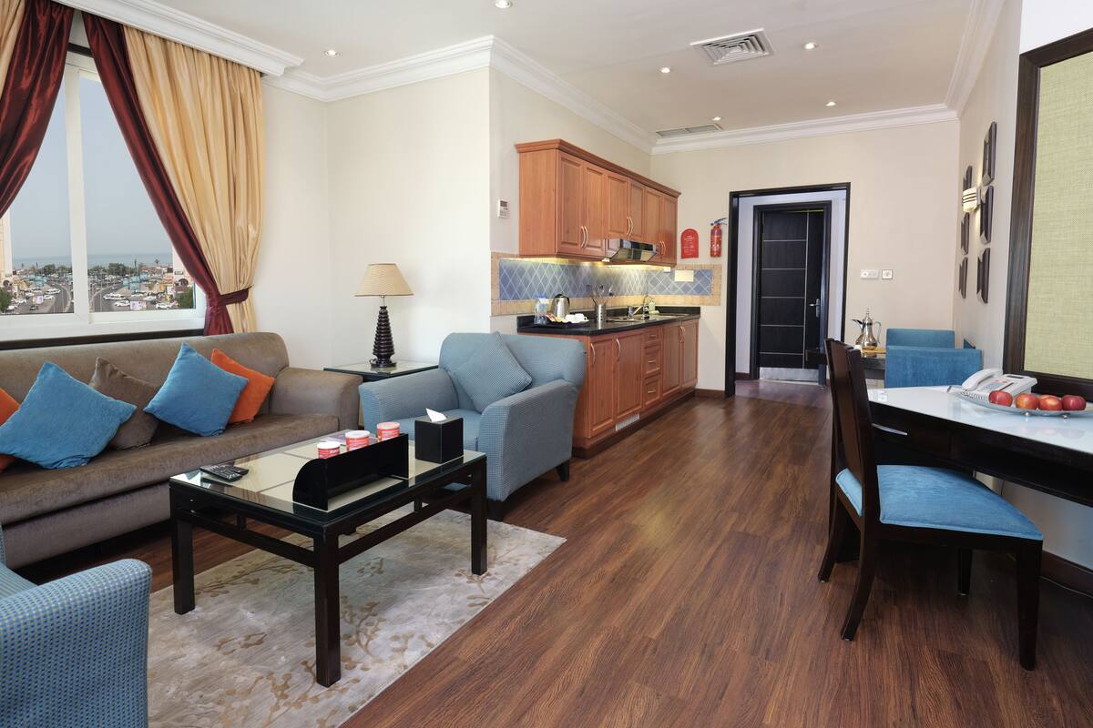 A modern living room with a sofa, armchairs, and a coffee table. There is a kitchen area with wooden cabinets and a dining table with chairs.