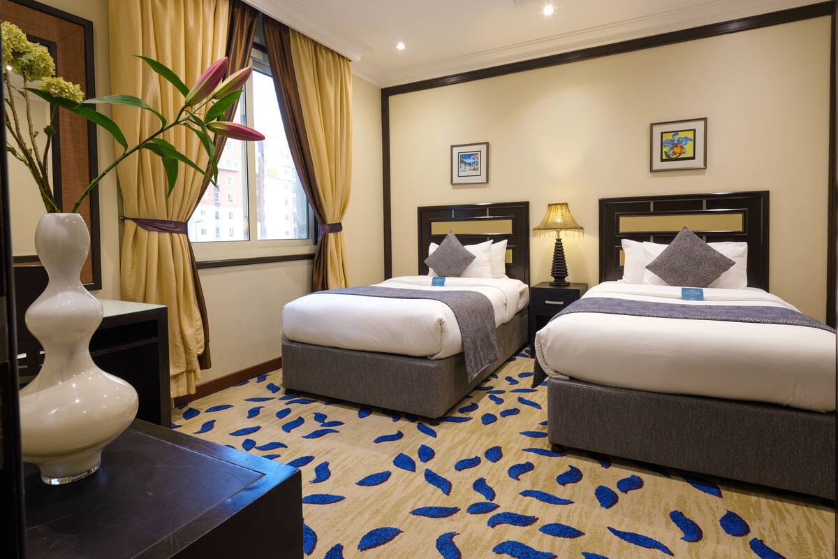 A hotel room with two beds, a nightstand with a lamp, a vase with flowers, and a window with curtains.