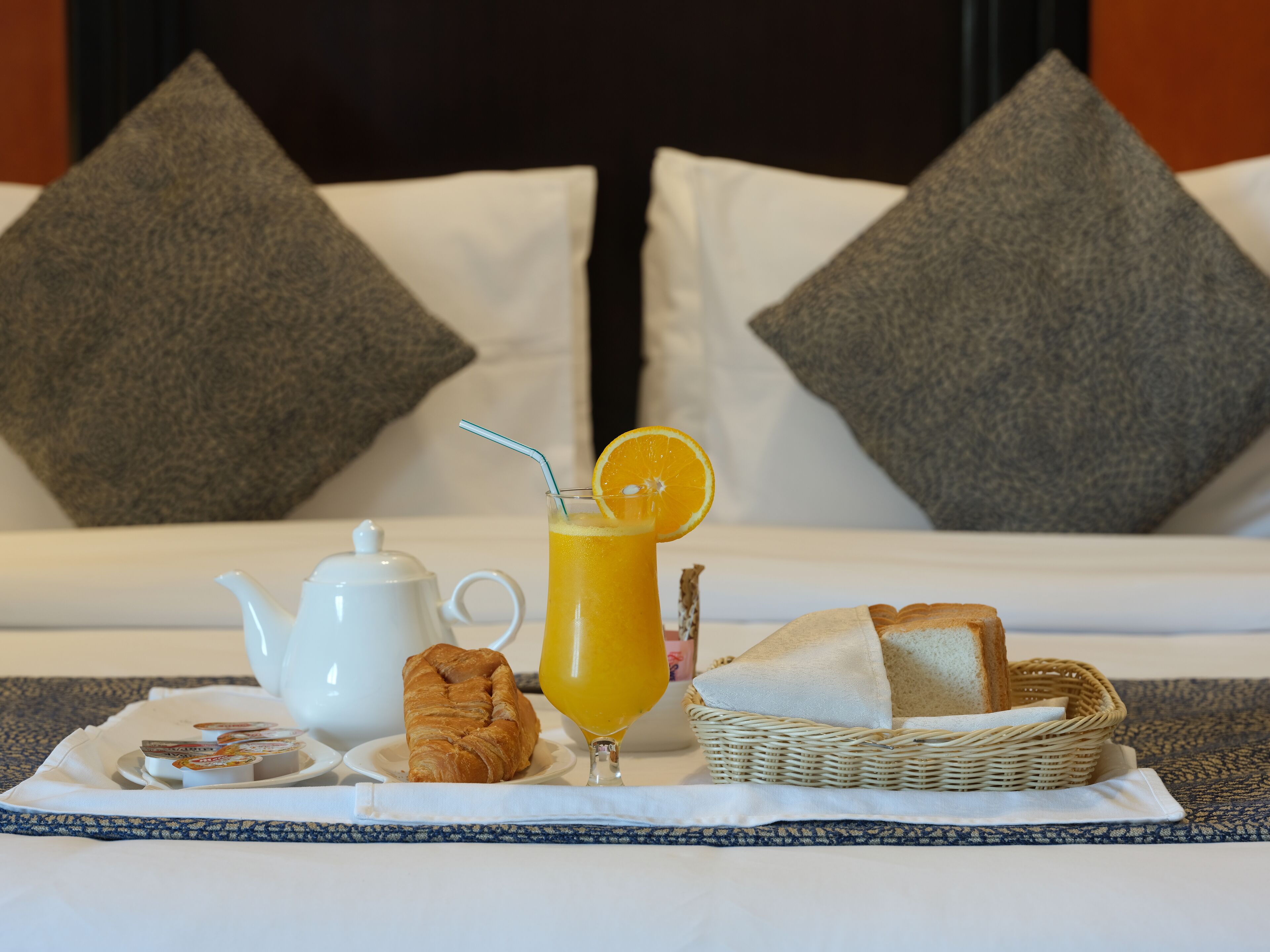 A breakfast setup on a bed with a teapot, orange juice, croissant, and bread basket.