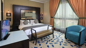 A hotel room with a large bed, a desk with a flat-screen TV, a chair, and a large window with curtains. - Boudl Kuwait (Salmiya)