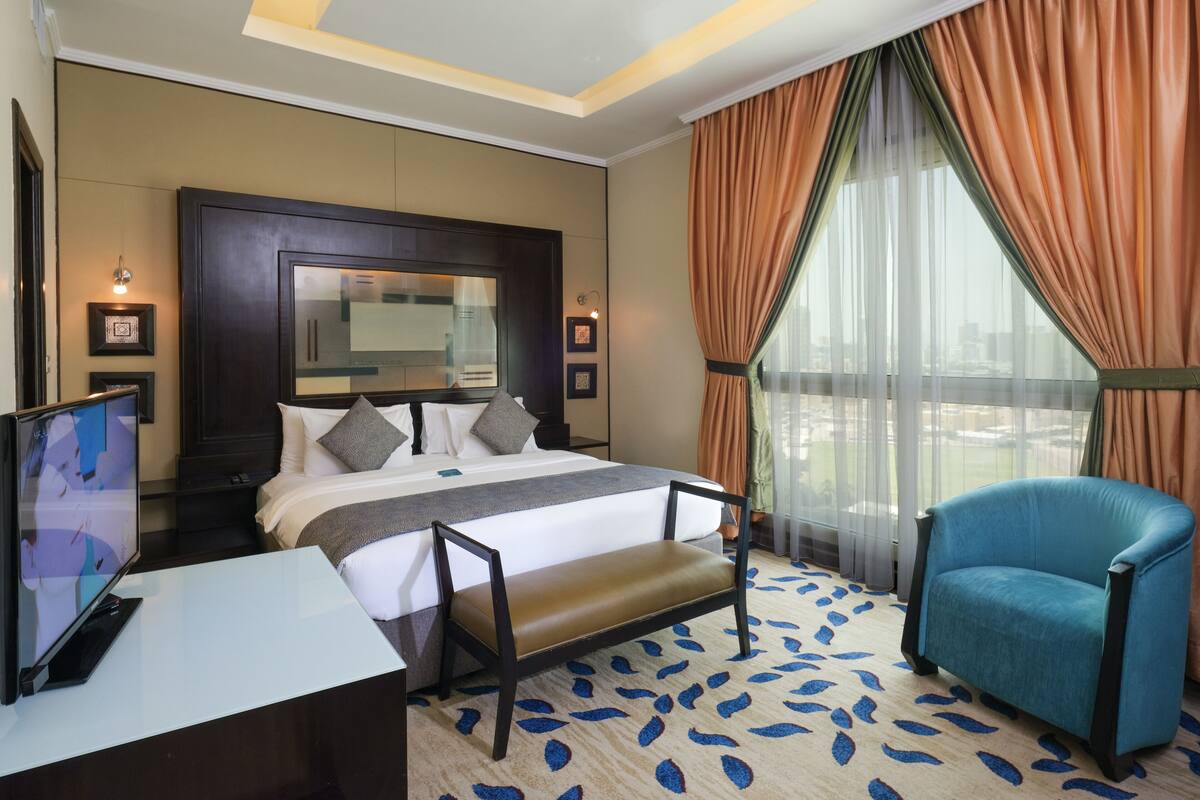 A hotel room with a large bed, a desk with a flat-screen TV, a chair, and a large window with curtains.
