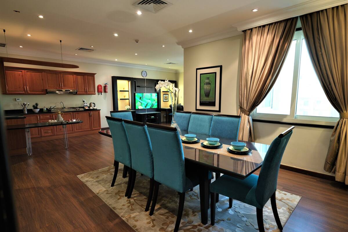 A modern kitchen with wooden cabinets, a dining area with blue chairs and a table, and a living area with a sofa and a TV.