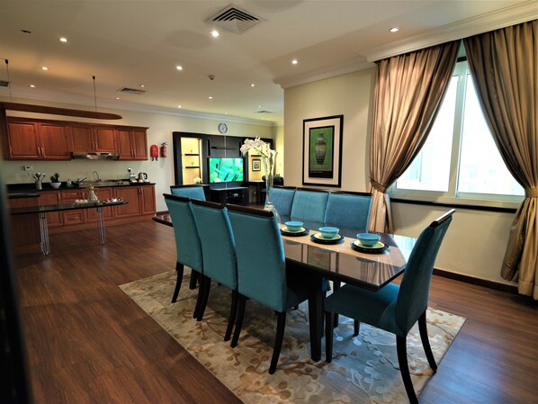 A modern kitchen with wooden cabinets, a dining area with blue chairs and a table, and a living area with a sofa and a TV. - Boudl Kuwait (Salmiya)