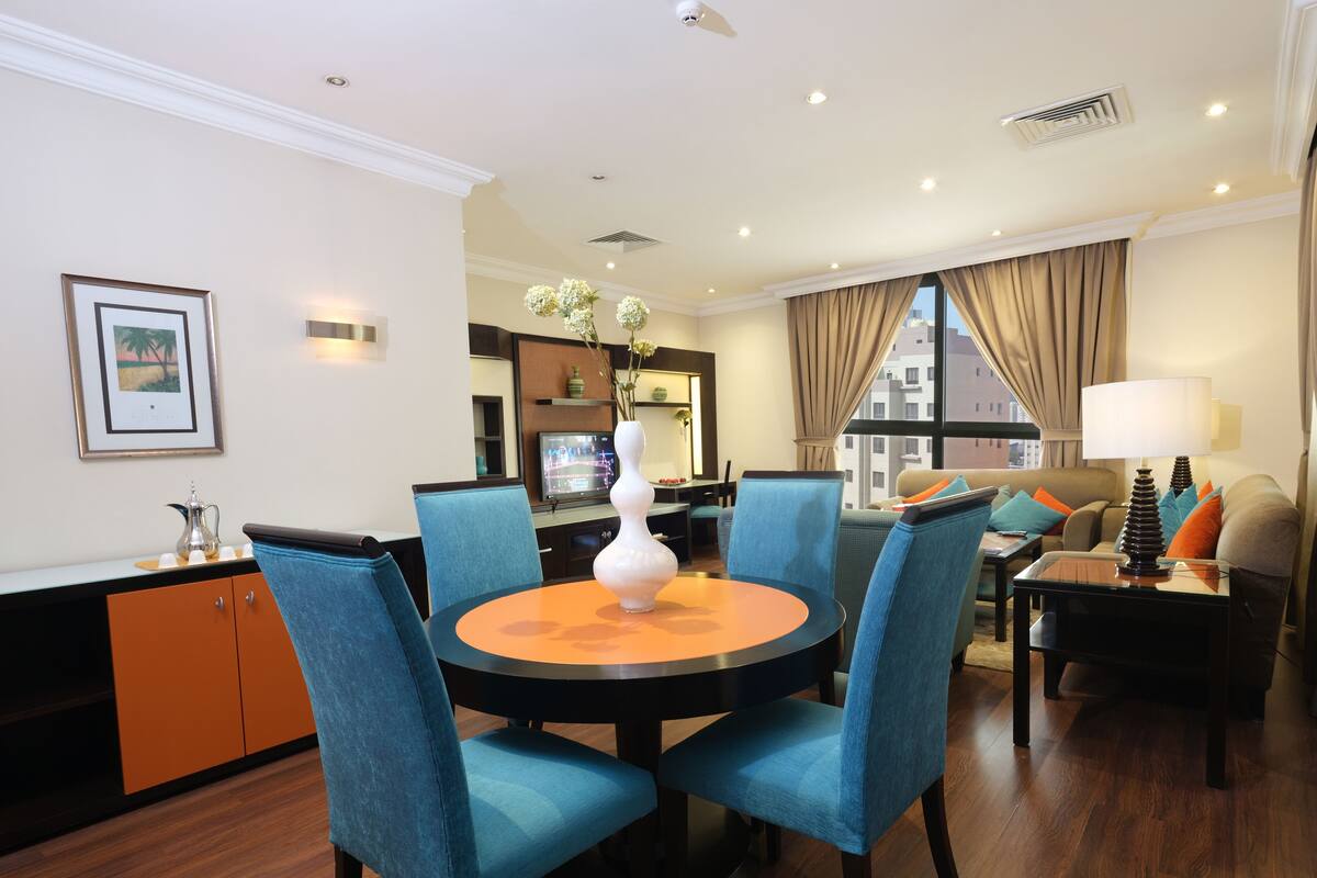A hotel room with a dining area, a sofa set, a TV, and a view of the city.