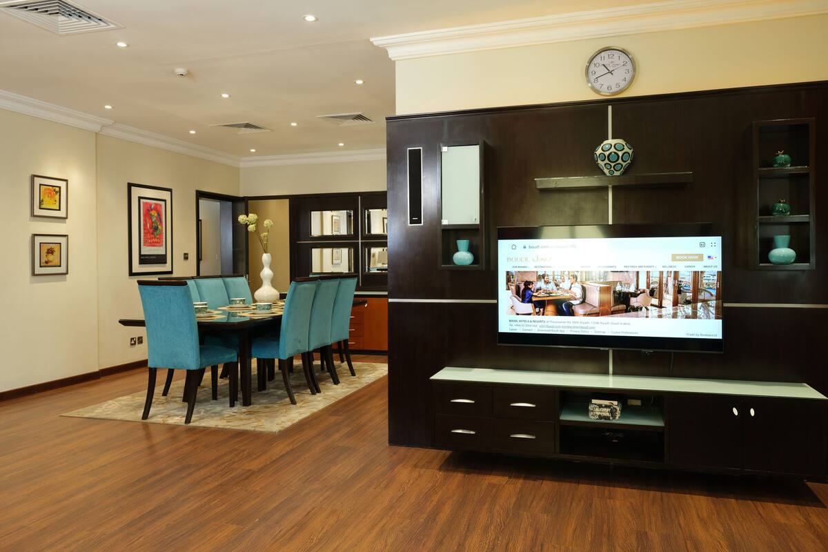 A modern living room with a dining area, a flat-screen TV mounted on a dark wood entertainment unit, and a clock on the wall.