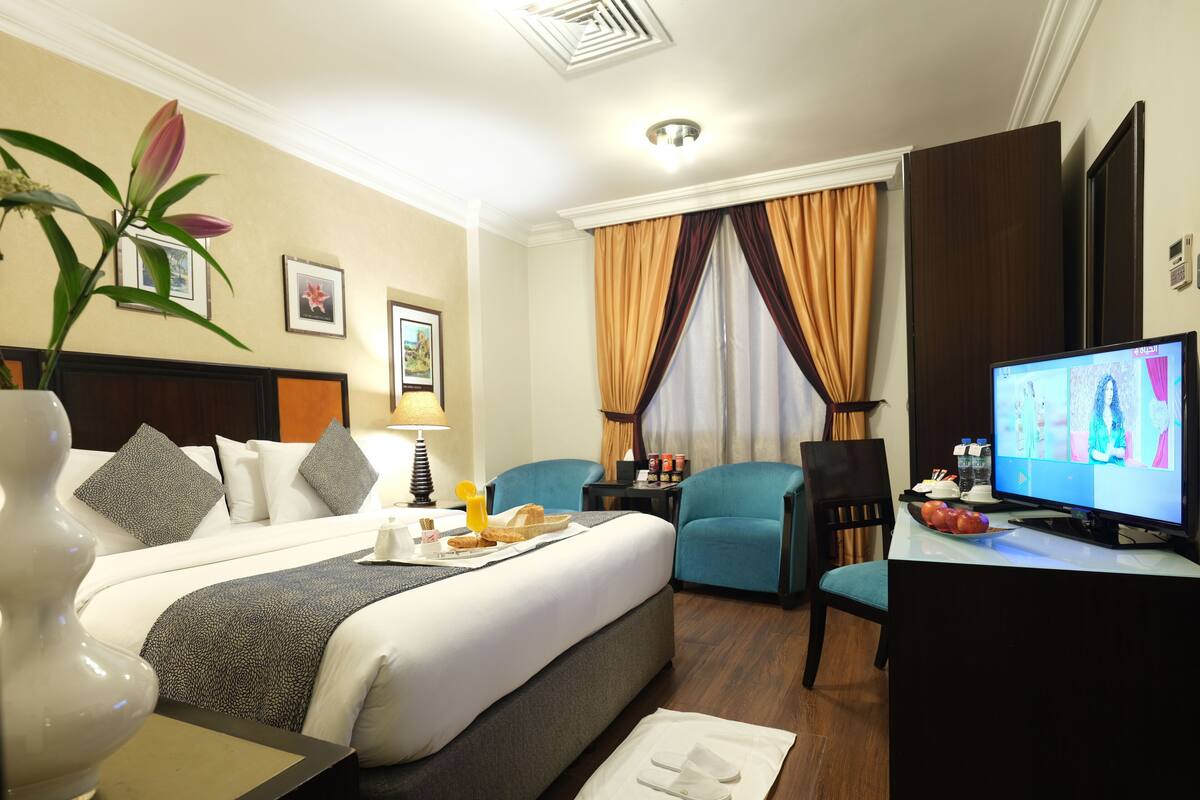 A hotel room with a bed, a desk with a computer, a chair, a lamp, and a plant.