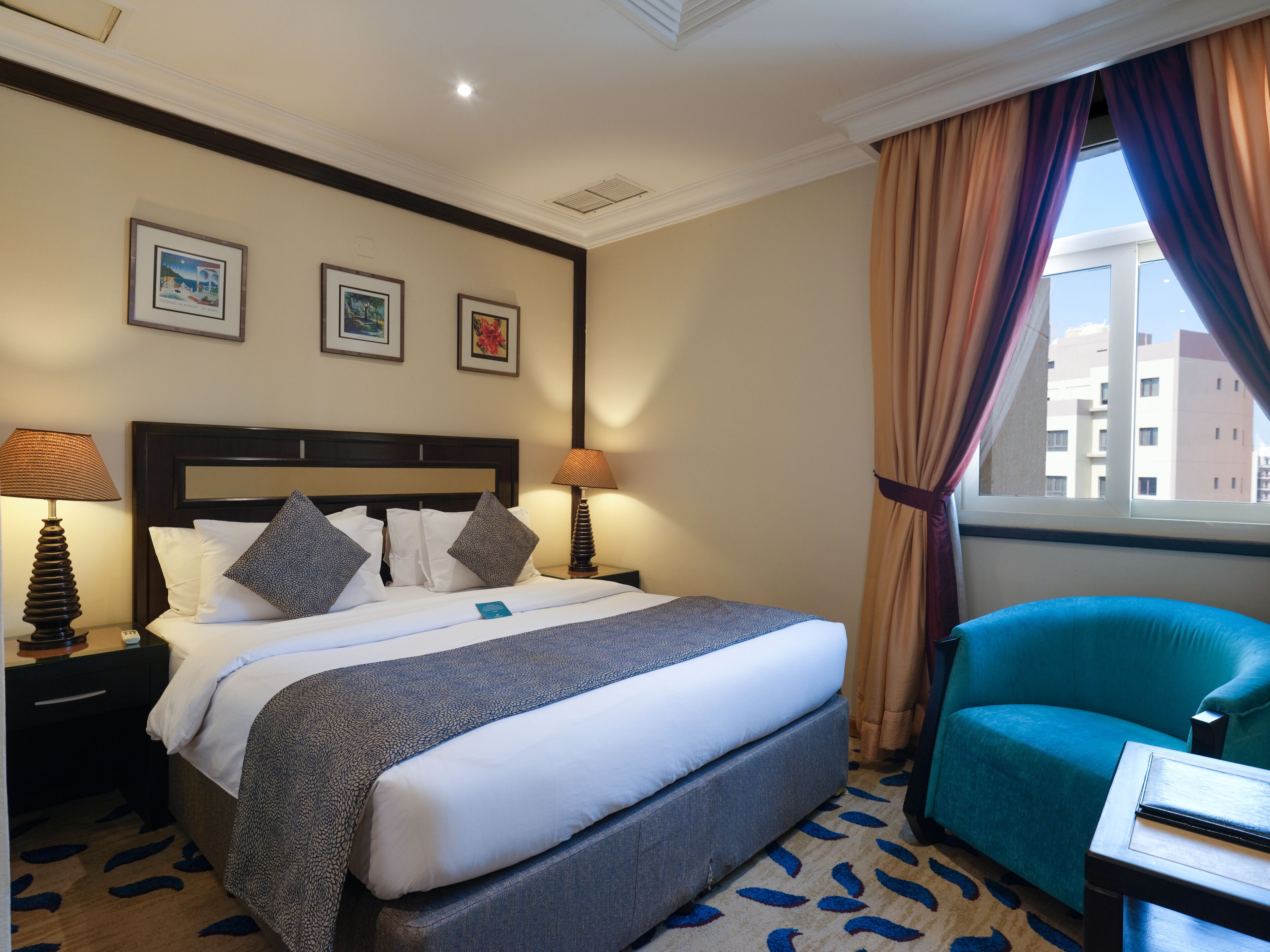 A hotel room with a bed, two bedside lamps, a nightstand, a blue armchair, and a window with curtains.