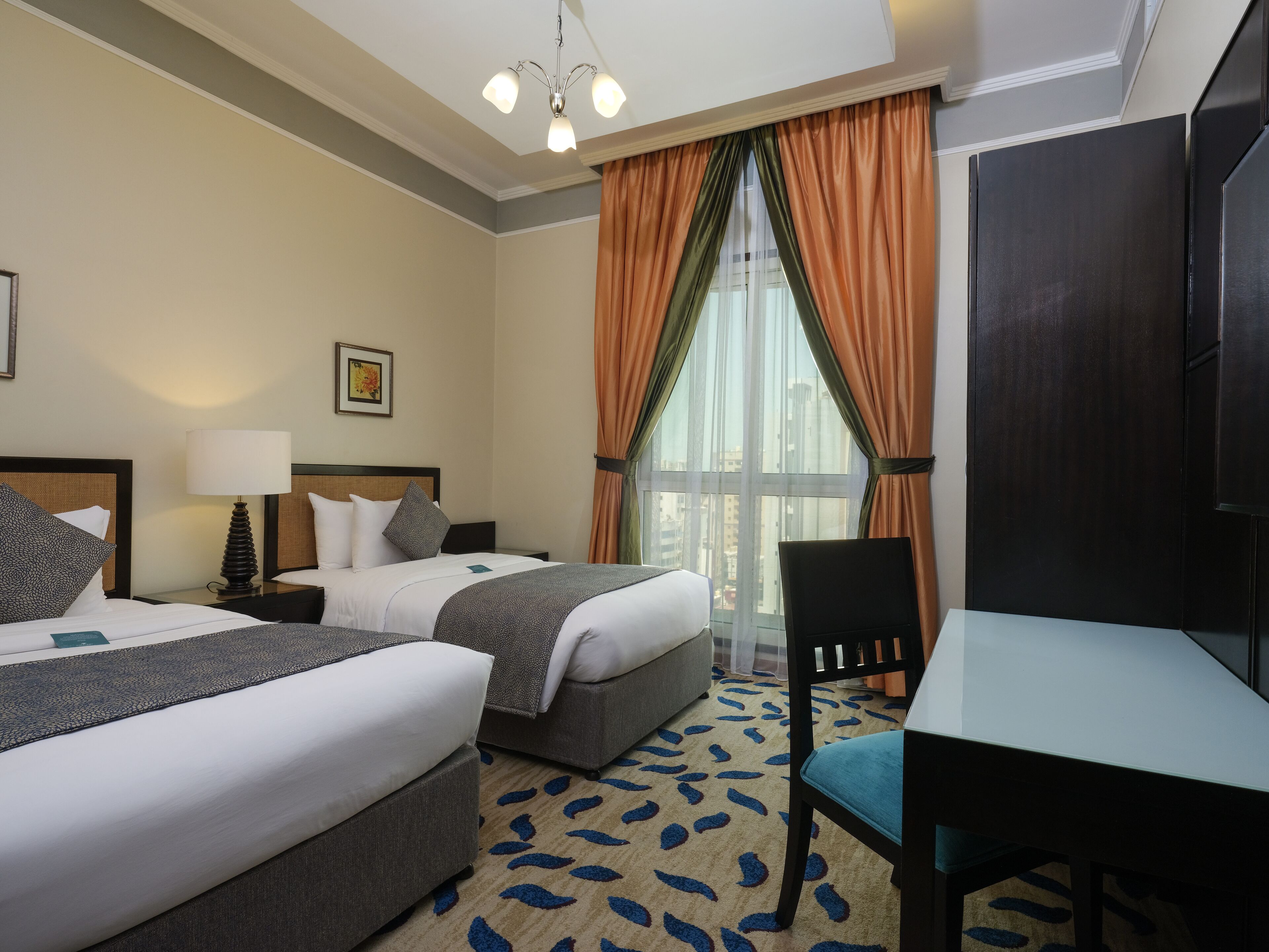 Une chambre d’hôtel avec deux lits, un bureau, une chaise, une lampe, un tableau au mur et une fenêtre avec des rideaux.