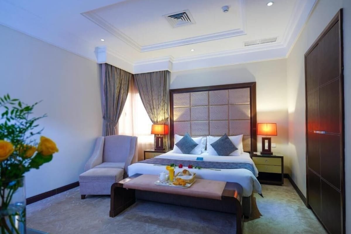 A hotel room with a bed, bedside tables, a chair, a bedside lamp, a nightstand, and a decorative vase with flowers.