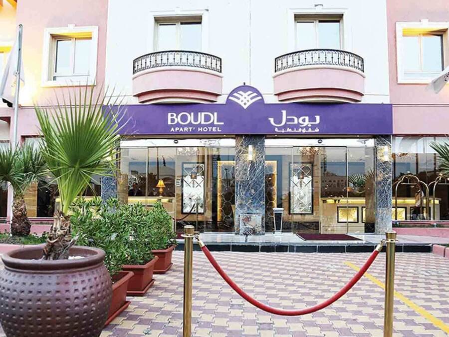 A hotel entrance with a sign reading "BOUDL APART HOTEL" and "BOUDL APART HOTEL" in another language. There are flags, potted plants, and a red rope barrier in front.