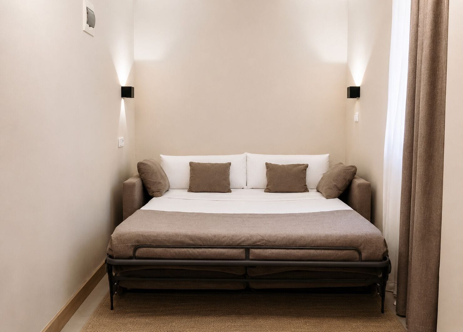 A bed with a headboard, two wall-mounted light fixtures, and a window with curtains.