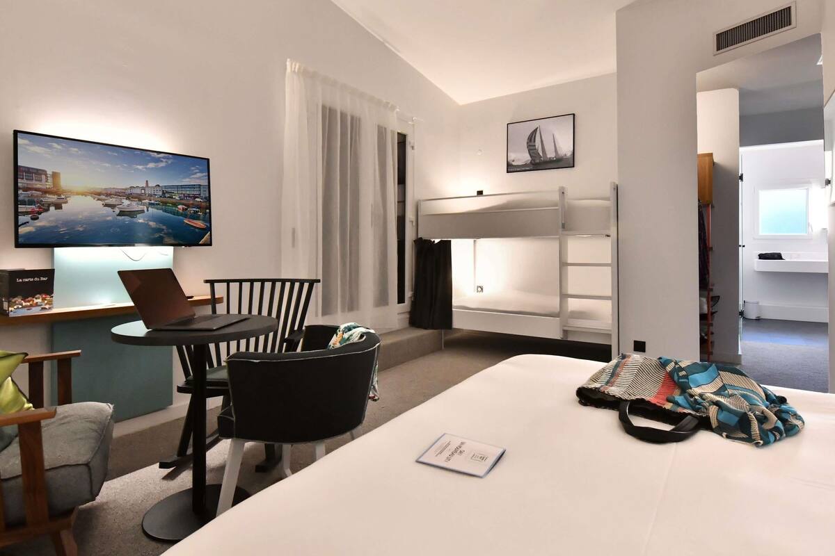 Habitación de hotel con una cama, un escritorio con una computadora portátil, una silla, una litera, un televisor y una ventana con cortinas.
