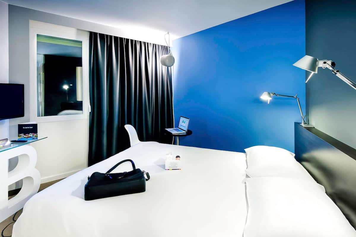 Habitación de hotel con cama, escritorio con laptop, silla, televisión y ventana con cortinas.