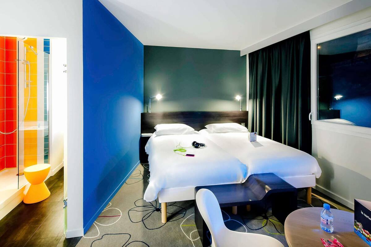Una habitación de hotel con cama, mesita de noche, silla y ventana con cortinas.