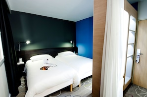 A hotel room with two beds, a dark blue accent wall, a wooden door, and a carpeted floor. - ibis Styles Brest Centre Port (Brest)